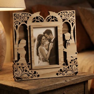 May include: Wooden photo frame with a laser-cut design featuring silhouettes of a bride and groom, trees, and floral accents. The frame holds a black and white photograph of a couple. The frame is displayed on a wooden surface.