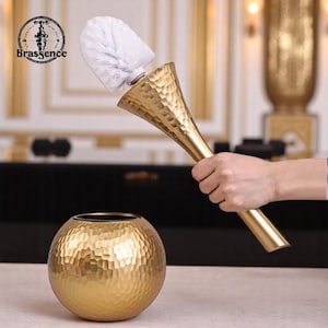 May include: A gold-coloured toilet brush with a textured handle and white bristles. The brush is held above a matching gold, textured, spherical holder. The word "Brassence" is printed on the top left of the image.