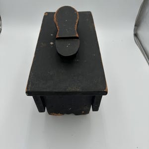 Puede incluir: Una caja de betún vintage, pintada de negro, con un asa de madera en forma de zapato. La caja tiene una base rectangular y probablemente está hecha de madera. La pintura muestra signos de desgaste, dándole un aspecto rústico.