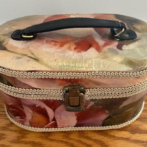 Pode incluir: Caixa oval vintage com um design floral. A caixa apresenta uma alça preta, um fecho de metal e é adornada com uma corda dourada. O padrão floral inclui tons de rosa, vermelho e creme.