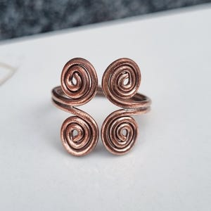 Puede incluir: Un anillo de metal color cobre con un diseño abierto que presenta cuatro espirales de alambre enrollado dispuestas en un patrón simétrico. El metal tiene un acabado oxidado oscuro en las hendiduras, resaltando la estructura del alambre.