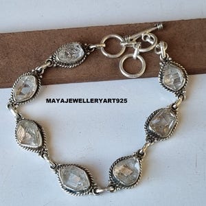 Puede incluir: Una pulsera de plata con piedras preciosas claras y facetadas engastadas en un diseño de bisel. La pulsera tiene un cierre de palanca y se muestra sobre una superficie marrón. El texto "MAYAEWELLERYART925" es visible.