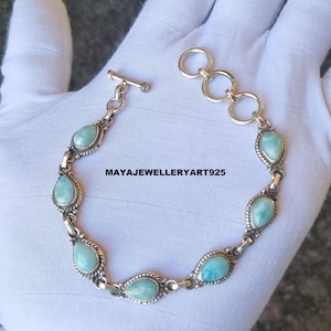May include: A silver bracelet featuring oval and teardrop-shaped, light blue gemstones. The bracelet has a toggle clasp and is connected by small, decorative links. The text "MAYAJEWELLERYART925" is visible on the bracelet.