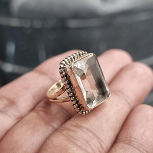 Puede incluir: Un anillo de tono dorado con una gran piedra preciosa rectangular, transparente y facetada, engastada en un marco con un borde texturizado de cuentas. La banda del anillo se divide en dos hilos metálicos dorados, finos, lisos y pulidos.