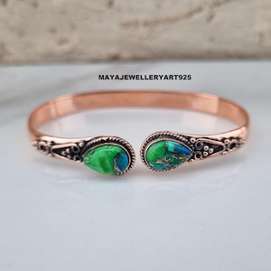 May include: Copper cuff bracelet with two teardrop-shaped green and blue stone accents. The bracelet has decorative detailing near the stone settings and the text "MAYAJEWELLERYART925".