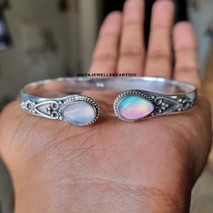 Peut inclure: Un bracelet manchette ouvert argenté orné de deux pierres d'opale en forme de poire. Les opales présentent des couleurs irisées. Le bracelet a des détails décoratifs autour des pierres. Le bracelet est présenté sur une main.