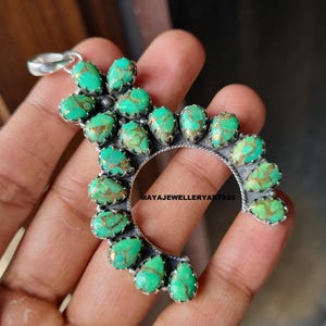 May include: A silver pendant featuring a crescent design, embellished with multiple teardrop-shaped turquoise stones with gold detailing. The pendant is held in a hand, highlighting the craftsmanship of the jewellery.