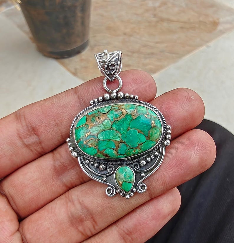 May include: A silver pendant with an oval, green stone with copper-colored inclusions. The stone is set in a detailed silver frame with a smaller, teardrop-shaped stone below. The pendant has a decorative bail for a chain.