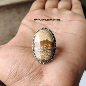 May include: An oval-shaped ring featuring a large, patterned stone in shades of beige and brown, set in a silver-colored metal band. The ring's stone has a unique, natural pattern. The ring is displayed on a hand. The text "MAYAJEWELLERYART925" is visible.
