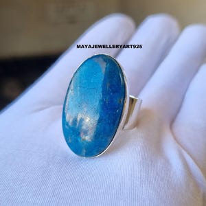 Puede incluir: Un anillo de plata con una gran piedra preciosa azul de forma ovalada. La piedra preciosa tiene una apariencia texturizada con diferentes tonos de azul. El anillo tiene una banda de plata ancha y sencilla. El texto "MAYAJEWELLERYART925" es visible.