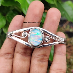 May include: A silver bracelet with an oval opal gemstone at the center. The opal displays a play of color with iridescent hues. The bracelet has a decorative design with swirls and is held by a hand. The text "MAYAJEWELLERYART925" is visible.