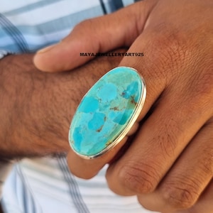 Könnte beinhalten: Ein großer ovaler Türkis-Ring in einer polierten Silberfassung. Der Stein zeigt eine leuchtende blau-grüne Farbe mit natürlichen braunen Matrix-Adern. Der Text MAYAJEWELLERYART925 ist auf der Oberfläche des Steins zu sehen.