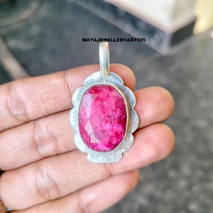 May include: A silver pendant featuring a large, oval-cut ruby gemstone. The ruby is a vivid pink-red hue, set within a silver bezel. The pendant has a decorative, scalloped border and a small loop for a chain, suitable for necklaces.