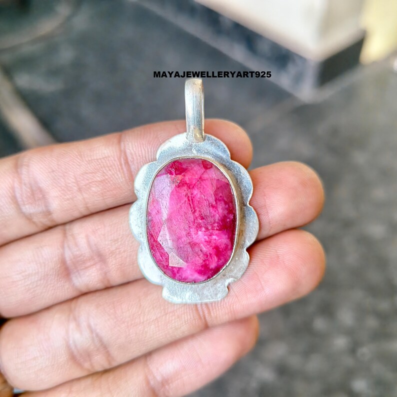 May include: A silver pendant featuring a large, oval-cut ruby gemstone. The ruby is a vivid pink-red hue, set within a silver bezel. The pendant has a decorative, scalloped border and a small loop for a chain, suitable for necklaces.