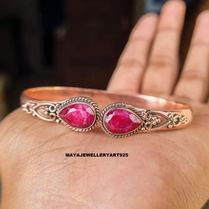 May include: A copper-colored cuff bracelet with two teardrop-shaped, vibrant red gemstones. The gemstones are set in a silver-colored metal with decorative detailing. The bracelet is open-ended and features the text "MAYAJEWELLERYART925".