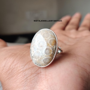 Pode incluir: Anel oval com uma pedra de coral fossilizado e uma banda de prata. O coral tem um padrão bege e branco. O anel está colocado numa mão, destacando o design e a textura da pedra. O texto "MAYAJEWELLERYART925" é visível.