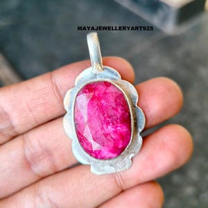 May include: A silver pendant featuring a large, oval-cut ruby gemstone. The ruby is a bright pink-red hue, set within a scalloped silver frame. The pendant has a small loop for a chain. Text on the image reads: MAYAJEWELLERYART925.