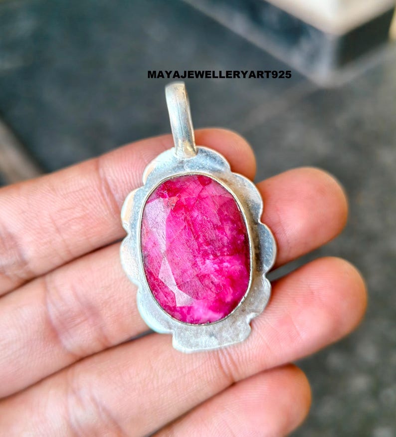 May include: A silver pendant featuring a large, oval-cut ruby gemstone. The ruby is a bright pink-red hue, set within a scalloped silver frame. The pendant has a small loop for a chain. Text on the image reads: MAYAJEWELLERYART925.
