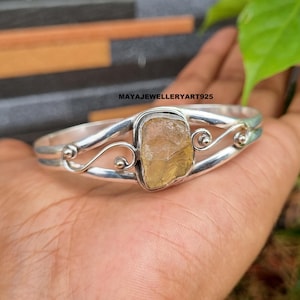 Op de afbeelding: Een zilverkleurige armband met een rechthoekige, lichtgele edelsteen in het midden. De armband heeft decoratieve krullen en een dubbelbandontwerp. De tekst "MAYAJEWELLERYART925" is zichtbaar.