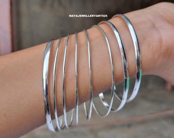 May include: A collection of shiny, silver bangles are stacked on a wrist. The bangles are thin and round, reflecting light. The jewelry is simple and elegant, suitable for everyday wear. The text "MAYAJEWELLERYART925" is visible.