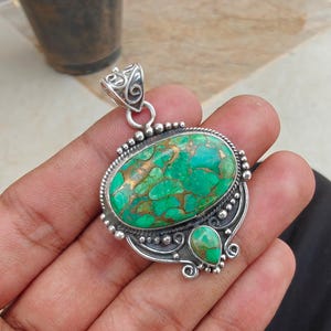 May include: A silver pendant with an oval, green and gold stone. The stone is surrounded by a silver frame with a decorative bail and a smaller, teardrop-shaped stone below. The pendant is held in a hand.