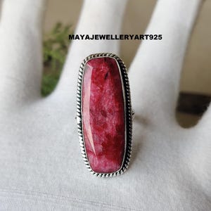 May include: A rectangular ruby ring with a silver-colored rope-like border. The ruby is a deep red color with lighter pinkish hues. The ring is set on a white surface. The text "MAYAJWELLERYART925" is visible.