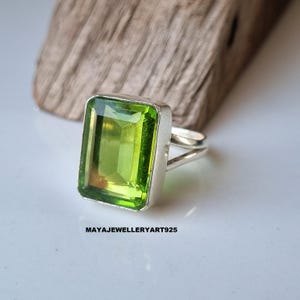 May include: A silver ring featuring a large, rectangular, light green gemstone. The gemstone has a vibrant, transparent appearance. The ring is set against a wooden background. The text "MAYAJEWELLERYART925" is visible.