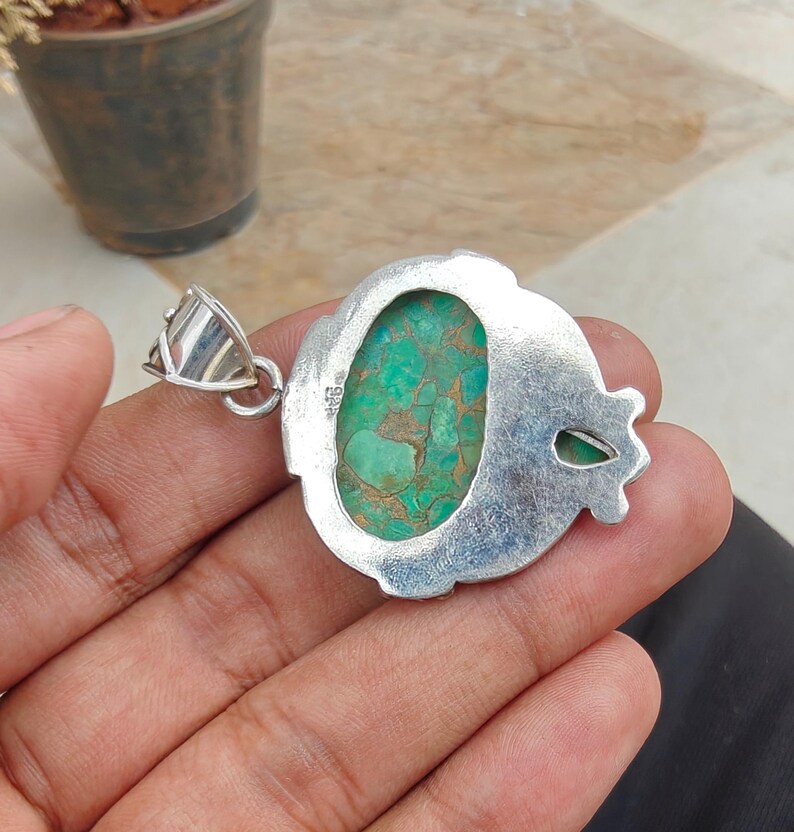 May include: A silver pendant with an oval, turquoise-colored stone. The stone has a mottled pattern of green and brown. The pendant has a decorative silver border and a silver bail for a chain.