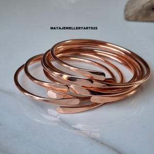 May include: A stack of copper-colored metal bangles. Each bangle has a smooth, rounded band that tapers to a flattened, open end. The jewelry is displayed on a light-colored surface. The text "MAYAJEWELLERYART925" is visible.