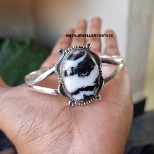 May include: A silver cuff bracelet featuring a large, oval zebra jasper stone with black and white patterns. The stone is set in a decorative frame. The bracelet has a simple, open design. Text on the image reads: MAYAJEWELLERYART925.