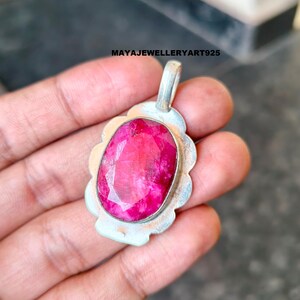 May include: A silver pendant featuring a large, oval-cut ruby gemstone. The ruby is a vibrant pink-red hue, set in a scalloped silver frame. The pendant has a loop for a chain. The text "MAYAJEWELLERYART925" is visible.