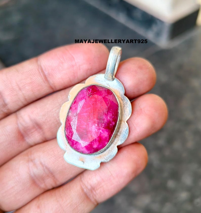 May include: A silver pendant featuring a large, oval-cut ruby gemstone. The ruby is a vibrant pink-red hue, set in a scalloped silver frame. The pendant has a loop for a chain. The text "MAYAJEWELLERYART925" is visible.