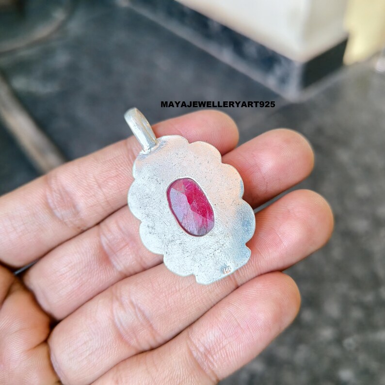 May include: A silver pendant featuring a central oval, deep red gemstone. The pendant has a scalloped edge and a loop for a chain. The text "MAYAJEWELLERYART925" is visible. The pendant is held in a hand.