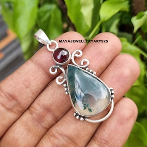 May include: A silver pendant with a teardrop-shaped moss agate stone and a small, round, red gemstone. The pendant has decorative swirls and the text "MAYAJEWELLERYART925". The pendant is held in a hand.