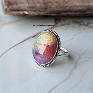 Pode incluir: Um anel de prata com uma pedra preciosa ovalada multicolorida. A pedra apresenta tons de amarelo, vermelho e roxo. O anel tem uma borda torcida em forma de corda ao redor da pedra e um design de haste dividida. O texto "MAYAJEWELLERYART925" é visível.