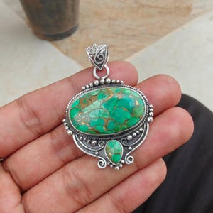 May include: A silver pendant with an oval, green and brown stone. The pendant has a decorative bail and a smaller, teardrop-shaped stone below the oval. The setting is detailed with small silver beads.