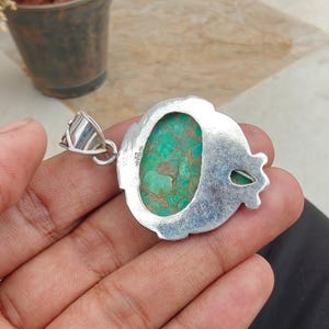 May include: A silver pendant with an oval, turquoise-colored stone. The stone has a mottled pattern of green and brown. The pendant has a decorative silver border and a silver bail for a chain.