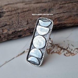 Puede incluir: Anillo de plata con diseño de fases lunares en una cara rectangular. El anillo tiene un borde detallado y el texto "MAYAJEWELLERYART925" es visible. Sobre un fondo de madera y mármol.
