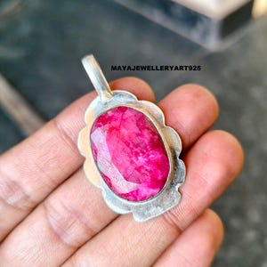 May include: A silver pendant featuring a large, oval-cut ruby gemstone. The ruby is a bright pink-red hue, set within a scalloped silver frame. The pendant includes a small loop for a chain. The text "MAYAJEWELLERYART925" is visible.