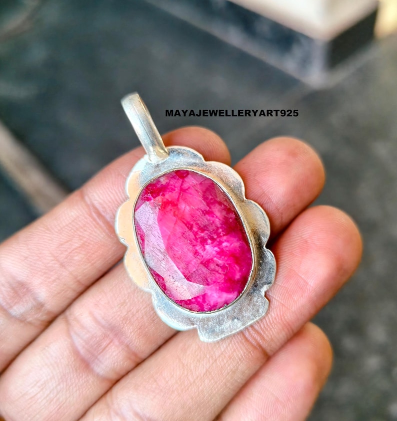 May include: A silver pendant featuring a large, oval-cut ruby gemstone. The ruby is a bright pink-red hue, set within a scalloped silver frame. The pendant includes a small loop for a chain. The text "MAYAJEWELLERYART925" is visible.