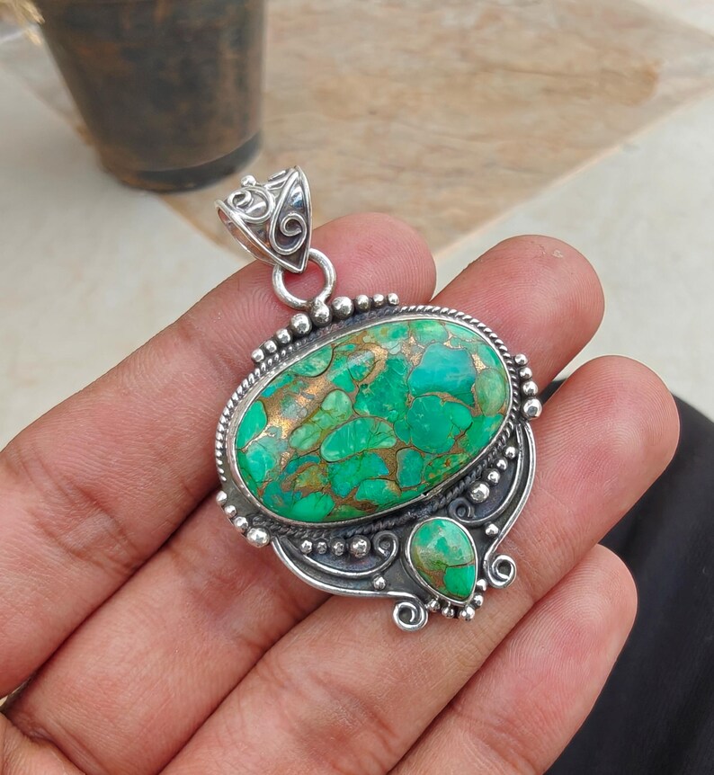 May include: A silver pendant with an oval, green and gold stone. The stone has a textured appearance and is surrounded by a silver frame with decorative beading. A smaller, teardrop-shaped stone is below the oval stone.