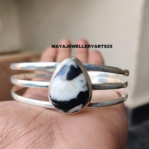 May include: A silver cuff bracelet with a teardrop-shaped stone. The stone features a black and white pattern. The bracelet has three parallel bands. The text "MAYAJEWELLERYART925" is visible.