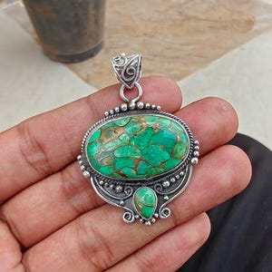 May include: A silver pendant with an oval, green stone with copper-colored inclusions. The stone is set in a detailed silver frame with a smaller, teardrop-shaped stone below. The pendant has a decorative bail for a chain.