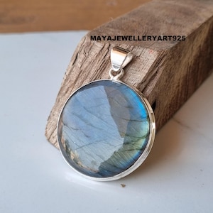 May include: A circular labradorite pendant with a silver bail. The stone has iridescent blue and green colours. The pendant is set in a silver bezel. The text "MAYAJEWELLERYART925" is visible on the wooden background.