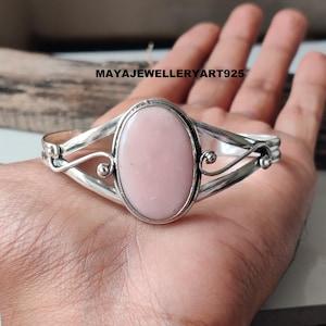 May include: A silver bracelet featuring a large, oval, pale pink stone. The bracelet has an open design with decorative swirls. The text "MAYAJEWELLERYART925" is visible. The bracelet is held in a hand.