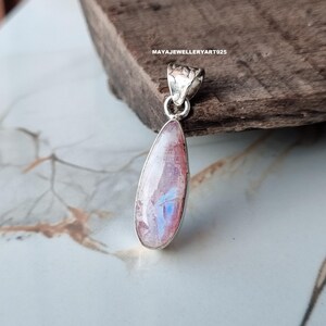 May include: A teardrop-shaped pendant with a polished, iridescent gemstone set in a silver bezel. The stone displays pink, white, and blue hues. The pendant has a decorative bail for a chain. The text "MAYAJEWELLERYART925" is visible.
