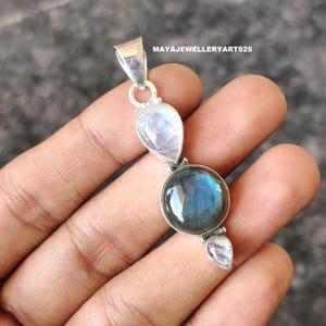 Puede incluir: Colgante de plata con tres piedras: una labradorita redonda e iridiscente y dos piedras lunares en forma de pera. El colgante tiene un lazo para una cadena y está marcado con el texto "MAYAJEWELLERYART925".