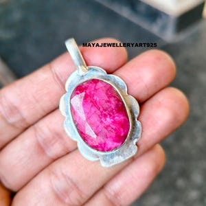 May include: A silver pendant featuring a large, faceted, oval ruby gemstone. The ruby is a bright pink hue, set in a decorative silver frame. The pendant has a loop for a chain. The text "MAYAJEWELLERYART925" is visible on the image.