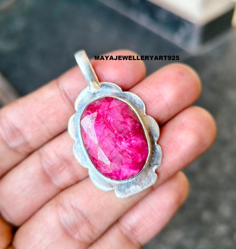 May include: A silver pendant featuring a large, faceted, oval ruby gemstone. The ruby is a bright pink hue, set in a decorative silver frame. The pendant has a loop for a chain. The text "MAYAJEWELLERYART925" is visible on the image.
