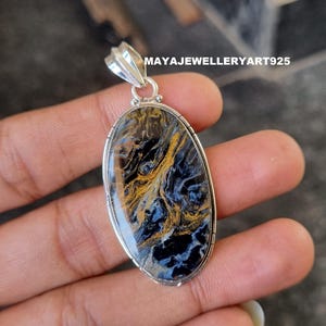 May include: Oval pendant with a silver bail. The gemstone has a swirling pattern of black, blue, and gold. The pendant is held in a hand. The text "MAYAJEWELLERYART925" is visible on the top right.